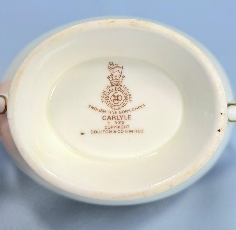 May include: The underside of a white bone china vessel featuring a brown stamp with a lion and crown emblem. Text reads: Royal Doulton, Made in England, English Fine Bone China, Carlyle, H. 5018, Copyright, Doulton & Co Limited. The base is oval.