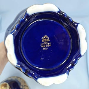 Może przedstawiać: Podstawa naczynia z głębokiego kobaltowego porcelany z falistym brzegiem i białym spodem. W centrum znajduje się złoty napis Lindner Kueps Bavaria Made in Western Germany Echt Cobalt 168 oraz mała ikona słońca nad nazwą marki.