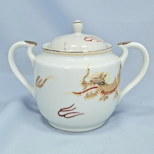 May include: White porcelain sugar bowl with a lid, decorated with a gold and red dragon motif. The bowl has two handles and gold trim. The dragon design is detailed, adding a touch of elegance to the piece.