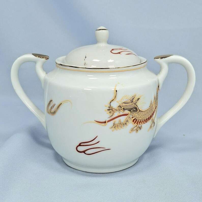 May include: White porcelain sugar bowl with a lid, decorated with a gold and red dragon motif. The bowl has two handles and gold trim. The dragon design is detailed, adding a touch of elegance to the piece.