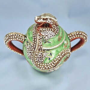 Pu&ograve; includere: Una teiera decorativa rotonda in ceramica verde con un motivo a drago. La teiera presenta un coperchio con una testa di drago e il corpo &egrave; adornato da un drago che lo avvolge. Le maniglie e il bordo sono marroni con accenti bianchi e dorati.