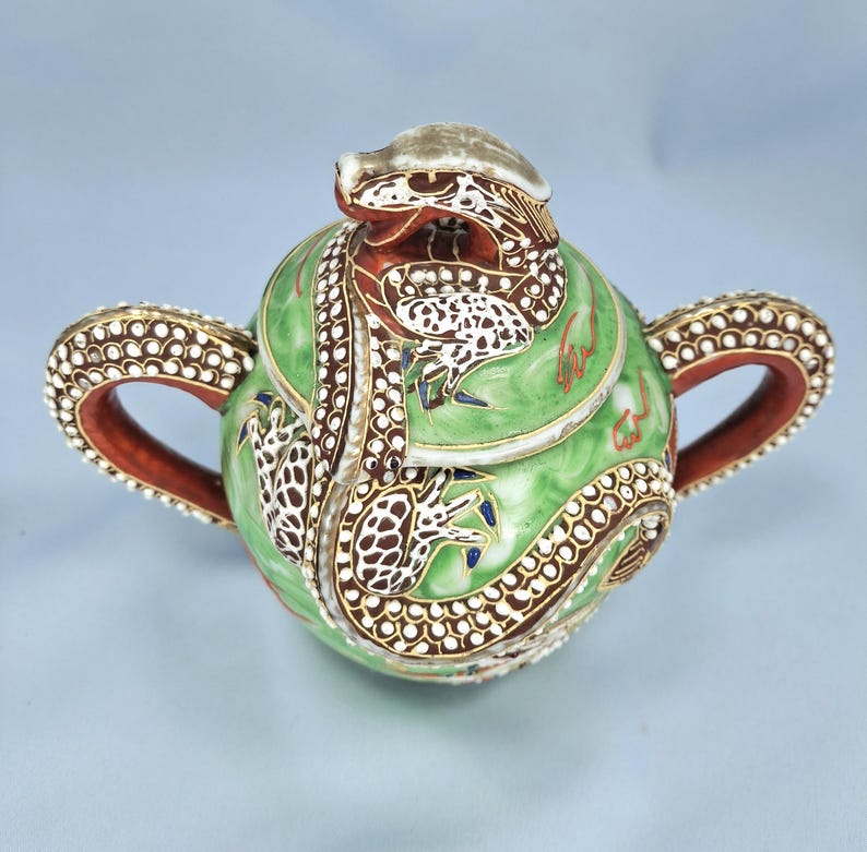 Pu&ograve; includere: Una teiera decorativa rotonda in ceramica verde con un motivo a drago. La teiera presenta un coperchio con una testa di drago e il corpo &egrave; adornato da un drago che lo avvolge. Le maniglie e il bordo sono marroni con accenti bianchi e dorati.