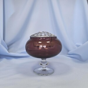 Puede incluir: Un jarr&oacute;n de flores de cristal morado oscuro vintage con una base clara y redondeada y una rejilla met&aacute;lica. El jarr&oacute;n tiene un cuerpo bulboso y un tallo corto, perfecto para arreglos florales. El fondo es una tela azul claro.