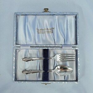 May include: A silver fork and spoon set inside a light blue box with a satin lining. The box is open, revealing the silverware and the text "Priestley & Moore Ltd CUTLERS & SILVERSMITHS SHEFFIELD - ENGLAND".
