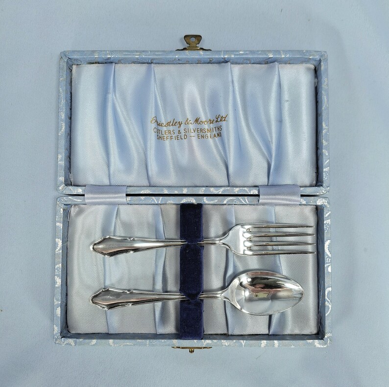 May include: A silver fork and spoon set inside a light blue box with a satin lining. The box is open, revealing the silverware and the text "Priestley & Moore Ltd CUTLERS & SILVERSMITHS SHEFFIELD - ENGLAND".