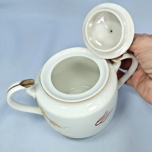 May include: White ceramic sugar bowl with a lid, featuring gold trim and decorative wheat and red accents. The bowl has two handles and a lid that is slightly ajar, revealing the interior. The design is classic and elegant.