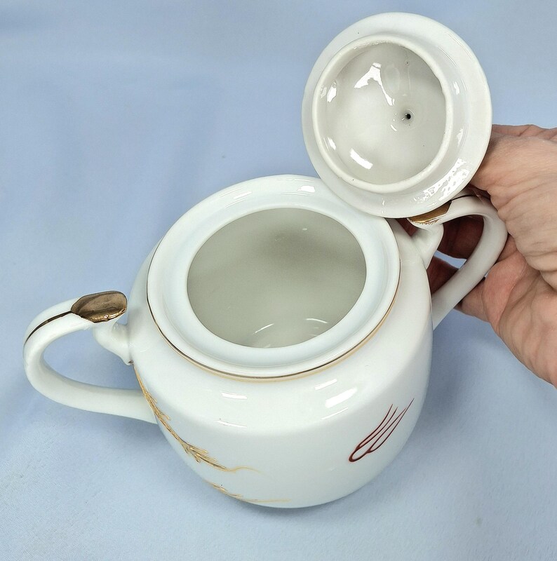 May include: White ceramic sugar bowl with a lid, featuring gold trim and decorative wheat and red accents. The bowl has two handles and a lid that is slightly ajar, revealing the interior. The design is classic and elegant.