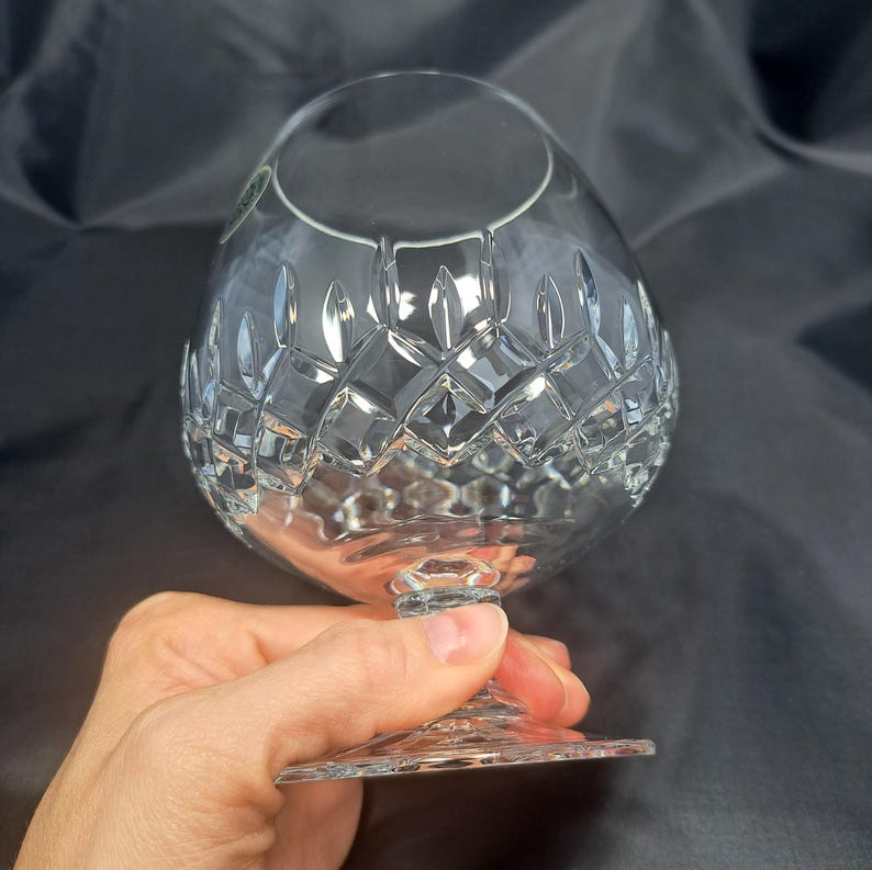May include: A clear crystal brandy snifter featuring a wide, rounded bowl with a detailed diamond-cut pattern around the midsection. The glass has a short, sturdy stem and a circular base, showcasing intricate geometric facets that reflect light.