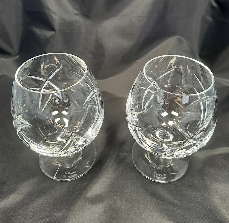 Puede incluir: Dos copas de brandy de cristal transparente con un patr&oacute;n geom&eacute;trico de estrella profundamente tallado en los cuencos. Cada copa tiene un tallo corto y una base circular con el texto grabado John Rocha WATERFORD en la parte inferior.
