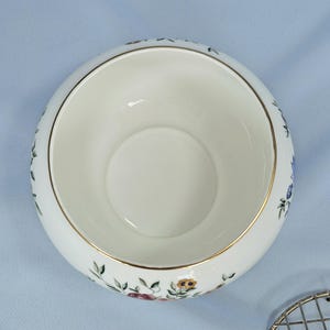 Puede incluir: Un cuenco de cer&aacute;mica blanco con borde dorado y estampado floral. El cuenco tiene forma redonda y una amplia abertura. El estampado floral incluye flores rojas, azules y amarillas con hojas verdes. El cuenco est&aacute; sobre un fondo azul claro.