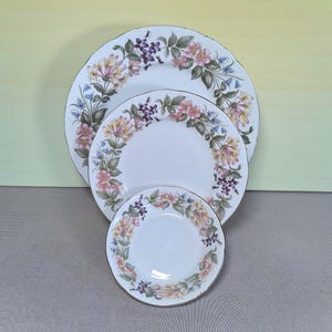 Puede incluir: Tres platos de cerámica blanca de diferentes tamaños, dispuestos uno dentro del otro. Cada plato presenta un diseño floral con flores rosas, amarillas y moradas, acentuado por hojas verdes y un borde dorado. Los platos están sobre un fondo neutro.