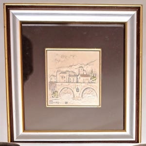 Puede incluir: Una obra de arte enmarcada que representa una ilustraci&oacute;n en blanco y negro de un peque&ntilde;o pueblo con un puente y &aacute;rboles. La obra est&aacute; en un marco plateado con un passe-partout marr&oacute;n.