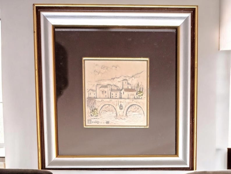 Puede incluir: Una obra de arte enmarcada que representa una ilustraci&oacute;n en blanco y negro de un peque&ntilde;o pueblo con un puente y &aacute;rboles. La obra est&aacute; en un marco plateado con un passe-partout marr&oacute;n.
