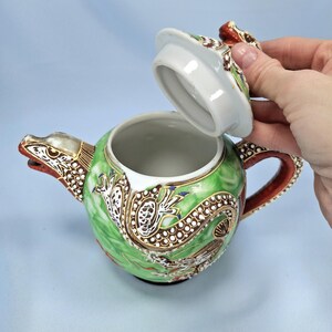 Pu&ograve; includere: Una teiera decorativa in ceramica con un design a drago. La teiera &egrave; principalmente verde con dettagli marroni, bianchi e rossi. Il coperchio &egrave; parzialmente aperto, rivelando l'interno. Il manico e il beccuccio sono finemente decorati.