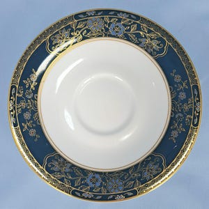May include: A round ceramic saucer featuring a plain white centre and a wide, dark blue border. The border is decorated with intricate gold-lined floral patterns and light blue flower accents, finished with a thin gold rim along the edge.