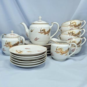 May include: A white porcelain tea set with gold dragon and red accent designs. The set includes a teapot, sugar bowl, creamer, cups, and saucers. The tea set is displayed on a light blue surface.