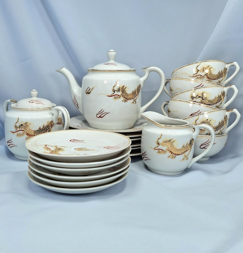 May include: A white porcelain tea set with gold dragon and red accent designs. The set includes a teapot, sugar bowl, creamer, cups, and saucers. The tea set is displayed on a light blue surface.