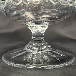 May include: A clear cut glass pedestal bowl featuring a faceted stem and a circular base with a radiating starburst pattern. The bowl has a scalloped rim and intricate geometric diamond cuts that reflect light across the transparent surface.