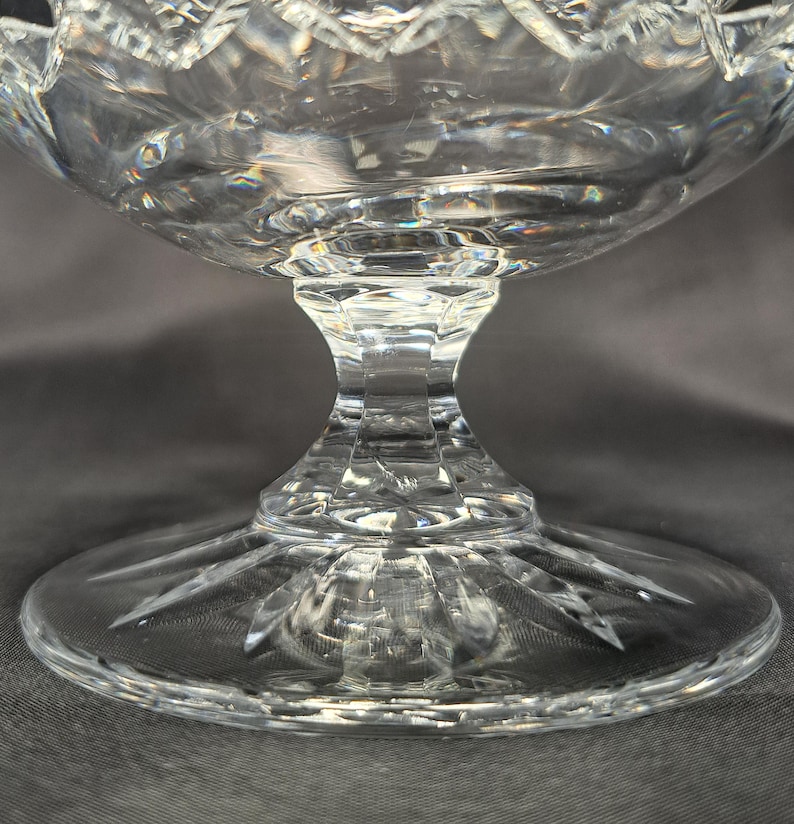 May include: A clear cut glass pedestal bowl featuring a faceted stem and a circular base with a radiating starburst pattern. The bowl has a scalloped rim and intricate geometric diamond cuts that reflect light across the transparent surface.