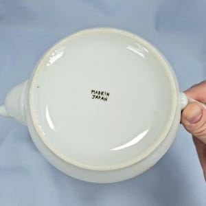 May include: White ceramic teapot with a spout and handle. The base of the teapot has the text "MADE IN JAPAN" in black lettering. The teapot is on a light blue surface.