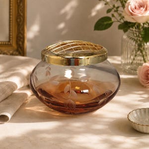 May include: A low, rounded glass bowl with a warm amber tint and delicate etched leaf patterns. The bowl features a removable, polished gold-tone metal grid lid designed for flower arranging, resting securely on the wide, circular glass opening.