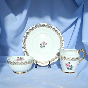 May include: A white porcelain tea set comprising a plate, bowl, and creamer. Each item features a gold filigree border and a pink rose design. The creamer has a gold handle and base. The set is presented against a blue backdrop.