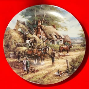 Pode incluir: Um prato de porcelana vintage com uma pintura de uma cena rural. A pintura representa uma carro&ccedil;a puxada por cavalos carregada de feno, dois cavalos, uma cabana com telhado de palha e v&aacute;rias figuras. A cena se passa em um campo com &aacute;rvores e uma vista distante de uma cidade.