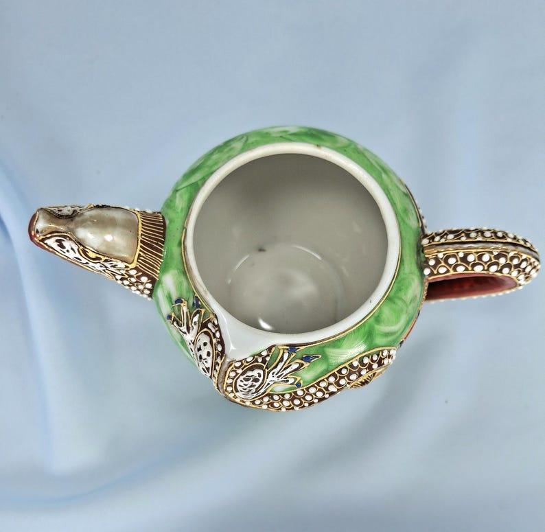 Pu&ograve; includere: Vista dall'alto di una teiera decorativa con un design verde, bianco e oro. La teiera ha un corpo rotondo, un beccuccio e un manico. Il manico e il beccuccio sono accentuati da dettagli dorati e bianchi. L'interno della teiera &egrave; bianco.