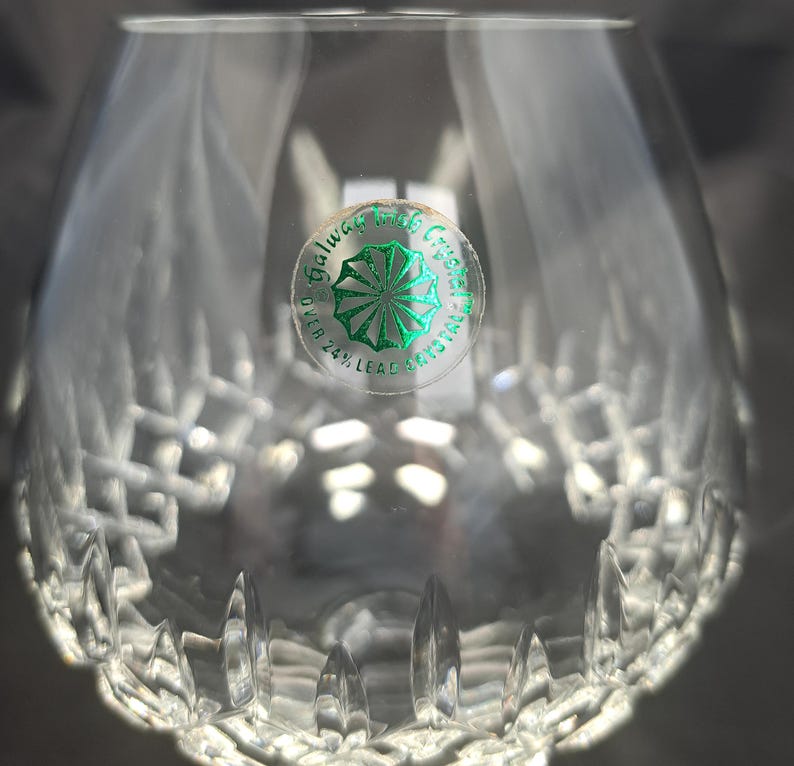 May include: A clear crystal brandy glass featuring a cut diamond pattern around the base and a circular green sticker on the side. The sticker displays the text Galway Irish Crystal and Over 24% Lead Crystal with a central starburst graphic.
