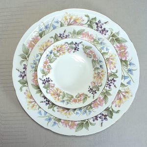 以下が含まれることがあります： 花柄の白い磁器のプレート3枚セット。プレートはスカラップエッジで、ピンク、黄色、紫の花、緑の葉、青いベル型の花で飾られています。プレートには金のトリムが付いています。