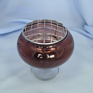 Puede incluir: Un cuenco redondo de cristal color ciruela con una base transparente con tallo. Una rejilla met&aacute;lica plateada cubre la parte superior del cuenco. El cristal tiene un acabado brillante, que refleja la luz. Esta pieza decorativa podr&iacute;a usarse para arreglos florales o como acento decorativo.
