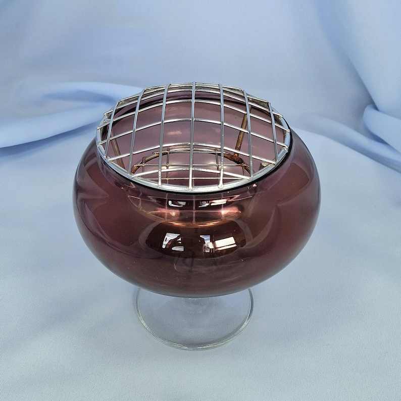 Puede incluir: Un cuenco redondo de cristal color ciruela con una base transparente con tallo. Una rejilla met&aacute;lica plateada cubre la parte superior del cuenco. El cristal tiene un acabado brillante, que refleja la luz. Esta pieza decorativa podr&iacute;a usarse para arreglos florales o como acento decorativo.
