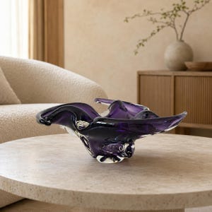 K&ouml;nnte beinhalten: Eine tiefviolette Glasschale mit unregelm&auml;&szlig;iger, organischer Form, flie&szlig;enden, wellenartigen R&auml;ndern und einer glatten, durchscheinenden Oberfl&auml;che. Die Schale hat einen dicken, klaren Glasboden, der die dunkelviolette, skulpturale Form st&uuml;tzt.