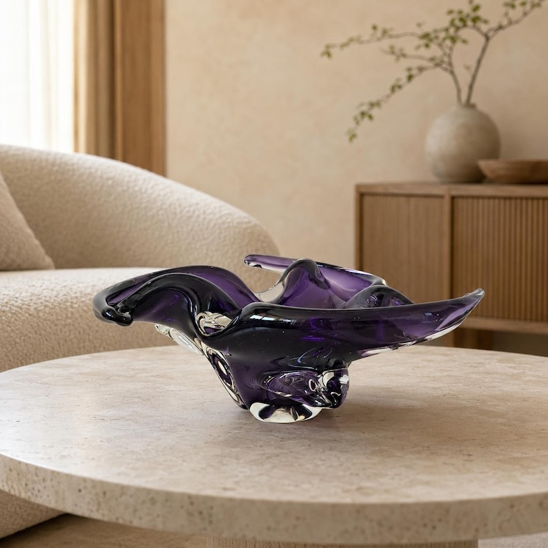 K&ouml;nnte beinhalten: Eine tiefviolette Glasschale mit unregelm&auml;&szlig;iger, organischer Form, flie&szlig;enden, wellenartigen R&auml;ndern und einer glatten, durchscheinenden Oberfl&auml;che. Die Schale hat einen dicken, klaren Glasboden, der die dunkelviolette, skulpturale Form st&uuml;tzt.