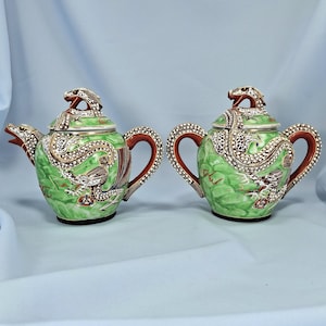 Pu&ograve; includere: Due teiere in ceramica verde abbinate con disegni di draghi. Le teiere hanno manici e beccucci marroni e i coperchi sono sormontati da figure di draghi. Le teiere sono decorate con accenti bianchi e marroni.