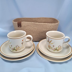 May include: Two off-white ceramic mugs and matching saucers featuring a botanical pattern of wheat, berries, and leaves with brown rim accents, positioned in front of an oval-shaped, woven tan rope basket with a small brown leather-like side tab.