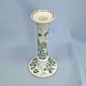 K&ouml;nnte beinhalten: Ein wei&szlig;er Kerzenhalter aus Keramik mit einem gr&uuml;nen Blumenmuster. Der Kerzenhalter hat einen runden Sockel, einen hohen, zylindrischen Schaft und eine flache Schale oben. Das Blumendesign umfasst Rosen und Bl&auml;tter.