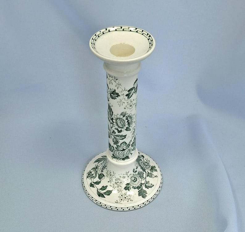 K&ouml;nnte beinhalten: Ein wei&szlig;er Kerzenhalter aus Keramik mit einem gr&uuml;nen Blumenmuster. Der Kerzenhalter hat einen runden Sockel, einen hohen, zylindrischen Schaft und eine flache Schale oben. Das Blumendesign umfasst Rosen und Bl&auml;tter.