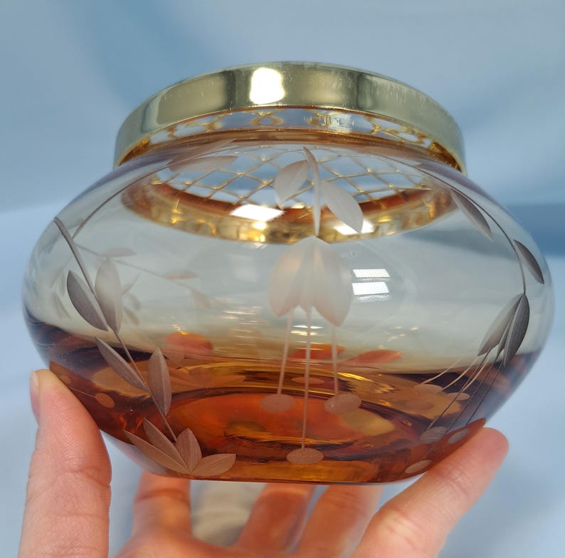 May include: A round amber glass bowl featuring a gold-tone metal rim and a gold-tone metal grid insert. The glass body is decorated with etched floral patterns, including leaves and stems, transitioning from a clear top to a deep amber base.