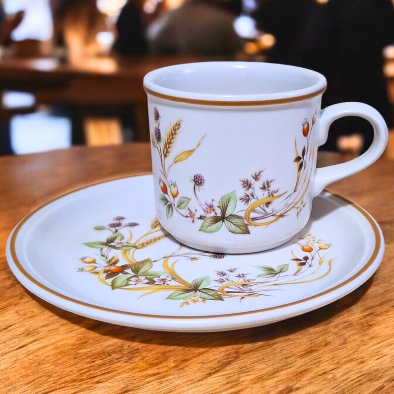 May include: A white teacup and saucer with a floral pattern featuring brown wheat stalks, green leaves, and red berries. The cup has a gold rim and a handle.