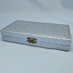 May include: A rectangular, light blue box with a floral pattern and a gold-colored clasp. The box has a decorative, embossed design on the lid and a gold trim around the edges. The box is closed and appears to be made of a sturdy material.