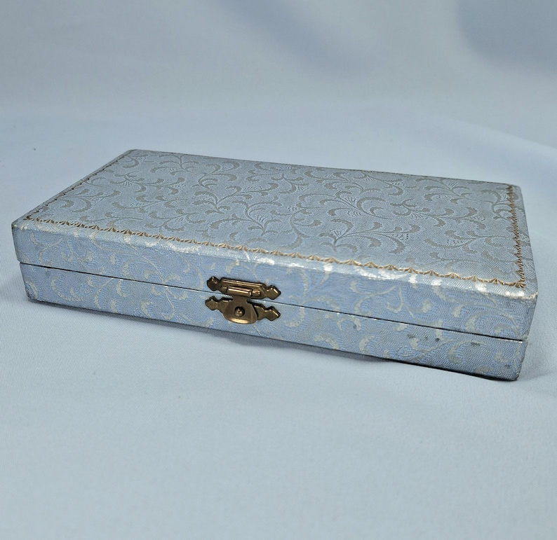 May include: A rectangular, light blue box with a floral pattern and a gold-colored clasp. The box has a decorative, embossed design on the lid and a gold trim around the edges. The box is closed and appears to be made of a sturdy material.