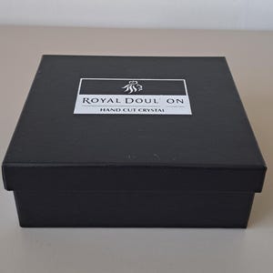 May include: A square black box with a lid featuring a white rectangular label on top. The label displays a lion crest icon above the text ROYAL DOULTON and the words HAND CUT CRYSTAL printed in black capital letters on a white background.