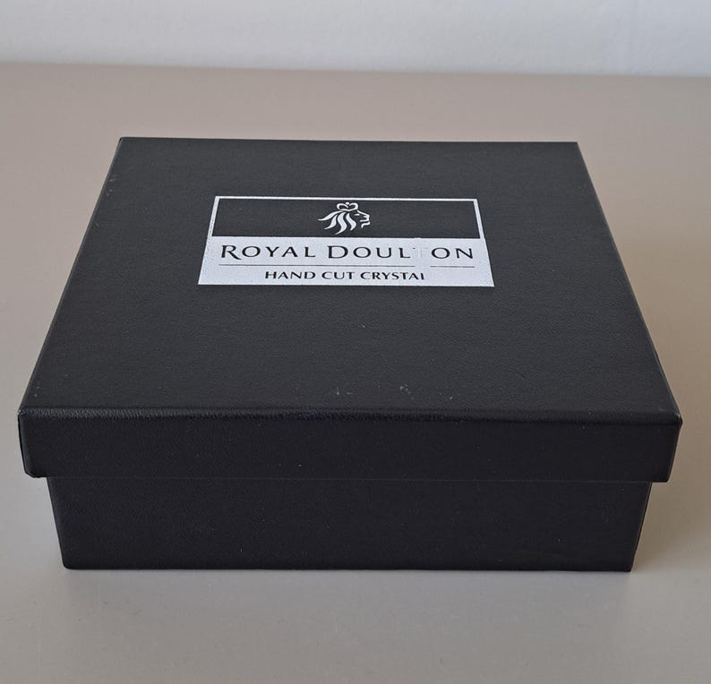May include: A square black box with a lid featuring a white rectangular label on top. The label displays a lion crest icon above the text ROYAL DOULTON and the words HAND CUT CRYSTAL printed in black capital letters on a white background.