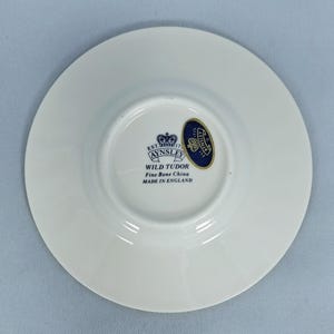 Peut inclure: Une soucoupe en porcelaine fine blanche avec un embl&egrave;me bleu et or. La soucoupe est de la collection Wild Tudor d'Aynsley, fabriqu&eacute;e en Angleterre. La soucoupe est ronde avec un centre l&eacute;g&egrave;rement sur&eacute;lev&eacute;.