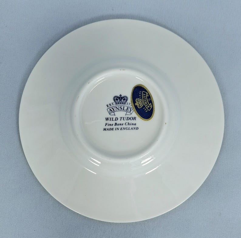 Peut inclure: Une soucoupe en porcelaine fine blanche avec un embl&egrave;me bleu et or. La soucoupe est de la collection Wild Tudor d'Aynsley, fabriqu&eacute;e en Angleterre. La soucoupe est ronde avec un centre l&eacute;g&egrave;rement sur&eacute;lev&eacute;.