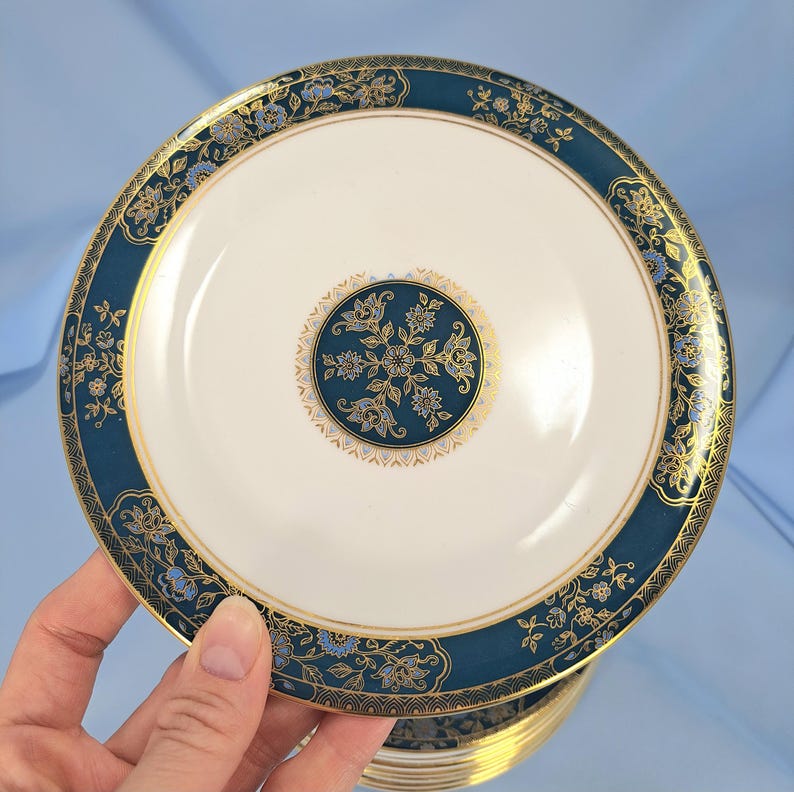 Op de afbeelding: Een rond keramieken bord met een cr&egrave;mekleurig midden en een centraal donkerblauwgroen medaillon versierd met gouden en lichtblauwe bloemmotieven. De brede rand is donkerblauwgroen met gouden filigraan, bloemmotieven en dunne gouden randen.