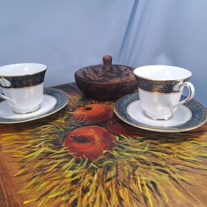 May include: Two white porcelain teacups with matching saucers featuring a dark blue and gold rim pattern, accompanied by a small round wooden lidded container, all arranged on a wooden surface painted with a depiction of red apples and hay.