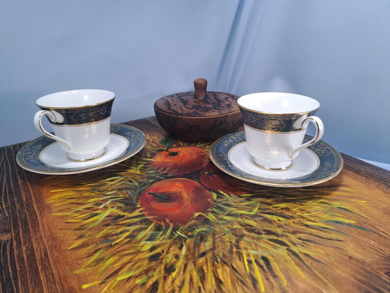 May include: Two white porcelain teacups with matching saucers featuring a dark blue and gold rim pattern, accompanied by a small round wooden lidded container, all arranged on a wooden surface painted with a depiction of red apples and hay.