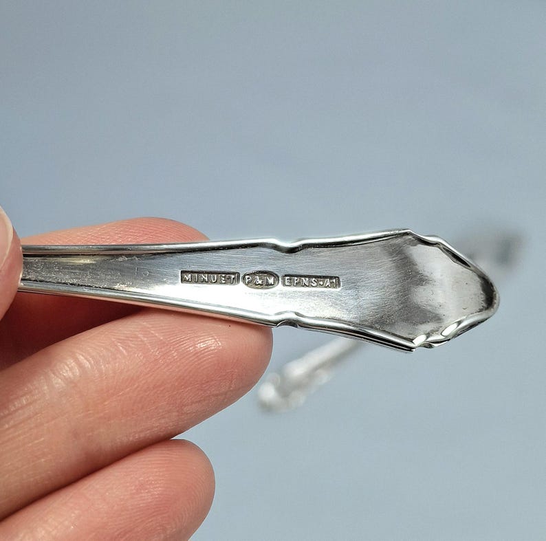 May include: Close-up of a silver spoon with the text "MINUET P&M EPNS-A1" stamped on the handle. The spoon has a decorative edge and is held against a light blue background.